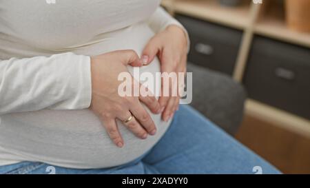 Junge hispanische schwangere Frau, die Herzform mit Händen am Bauch schafft, zuhause. Stockfoto
