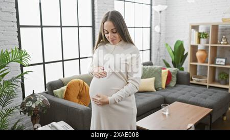 Eine junge hispanische schwangere Frau steht nachdenklich in einem gemütlichen Wohnzimmer und verkörpert Mutterschaft und Erwartung. Stockfoto