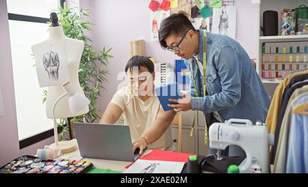 Zwei Männer arbeiten in einer Schneiderei zusammen, wobei einer einen Laptop benutzt und der andere ein Tablet hält, umgeben von Nähutensilien und Stoff. Stockfoto