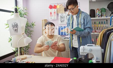 Zwei Männer arbeiten in einem Schneiderladen zusammen, der eine zählt Yuan und der andere macht Notizen zwischen Designskizzen und einer Schaufensterpuppe. Stockfoto