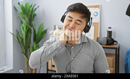 Ein junger asiatischer Mann mit Kopfhörern, der Nackenschmerzen in einem modernen Büro erlebt. Stockfoto