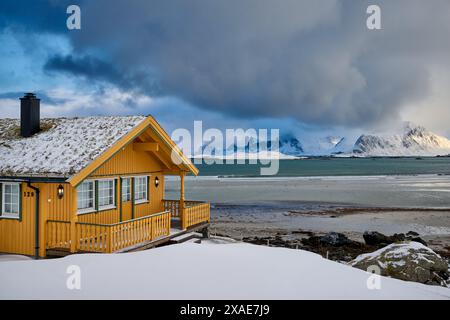 Gelbes Holzhaus oder Hütte in Winterlandschaft mit Meer und Bergen bei Sandbotnen, Lofoten, Norwegen, Europa Stockfoto