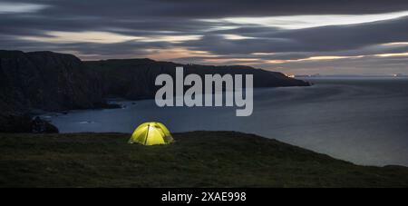 Panoramablick auf ein grünes beleuchtetes Campingzelt auf einer grasbewachsenen Klippe mit Blick auf das Meer in der Abenddämmerung. St. ABB's Head, Ostküste Schottlands. Wildes Camping Stockfoto