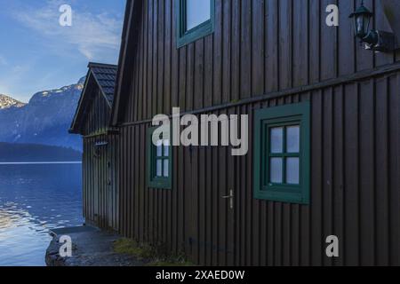 Fotografie eines dunkelbraunen Holzhauses am Alpensee von Hallstatt, umgeben von Bergen, am sonnigen Winterabend Stockfoto