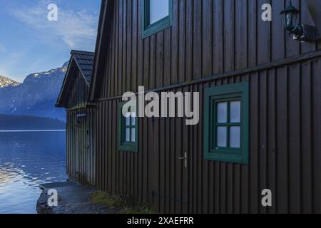 Fotografie eines dunkelbraunen Holzhauses am Alpensee von Hallstatt, umgeben von Bergen am sonnigen Winterabend Stockfoto