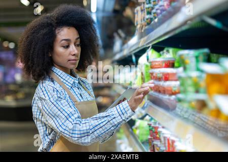 Junge weibliche Mitarbeiterin im Lebensmittelgeschäft, die mit einem Tablet den Lagerbestand in einem Gang mit verschiedenen Produkten verwaltet. Stockfoto
