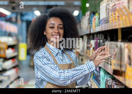 Eine fröhliche Arbeiterin trägt eine Schürze und arrangiert Gegenstände in einem Regal in einem hell beleuchteten Supermarktgang. Stockfoto