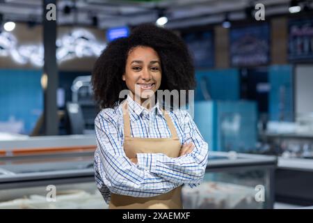 Selbstbewusste Frau mit überkreuzten Armen lächelnd in die Kamera, arbeitet auf einem Fischmarkt. Sie trägt ein kariertes Hemd und eine Schürze im Einzelhandel. Stockfoto