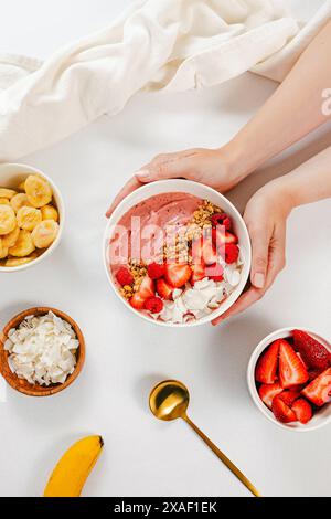 Weibliche Hände, die auf den Tisch legen Erdbeere Smoothie Schüssel mit Beeren, Müsli und Kokoschips, gesundes Frühstück Stockfoto
