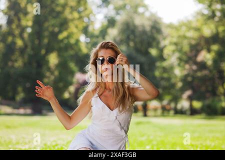 Eine selbstbewusste Frau in weißem Sommerkleid und Sonnenbrille posiert anmutig in einem üppig grünen Park und vermittelt eine entspannte und unbeschwerte Sommerstimmung. Stockfoto