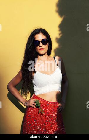 Elegante junge Frau mit Sonnenbrille und einem lebhaften Sommeroutfit vor einer kontrastierenden gelben Wand. Das Bild strahlt Stil und Selbstvertrauen aus. Stockfoto