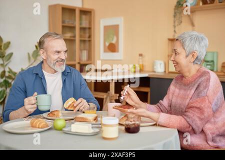 Porträt eines glücklichen Seniorenpaares, das zusammen in der gemütlichen Küche frühstückt und sich lächelnd ansieht Stockfoto