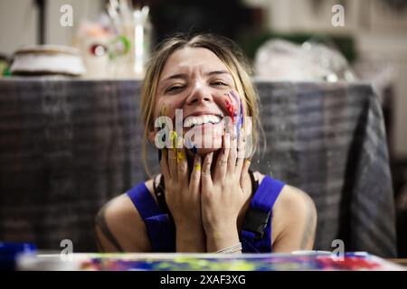 Eine junge Künstlerin drückt ihre Freude im Kunststudio aus, bedeckt mit bunten Farbspritzern auf ihren Händen und im Gesicht, während einer kreativen Sitzung. Stockfoto