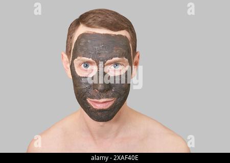 Schwarze Tonmaske auf dem Männergesicht. Kosmetologie Mann Porträt mit Schlamm gesundes Peeling. Kohle Natur Öko Masque. Dermatologisches Holzkohleprodukt. Häusliche Hautpflege. Hautpflege-Männerbehandlung. Grau Stockfoto