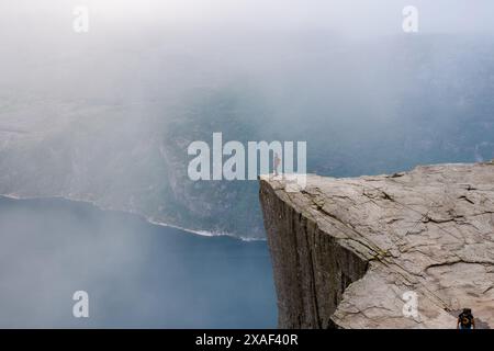 Preikestolen, Norwegen, Ein einsamer Wanderer steht auf dem Abgrund einer dramatischen Klippe in Norwegen, vor ihnen ziehen sich nebelige Land- und Wasserflächen. Stockfoto