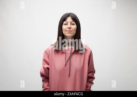 Eine ruhige und komponierte junge Frau trägt einen modischen rosa Hoodie und posiert selbstbewusst vor einem weichen grauen Hintergrund. Stockfoto