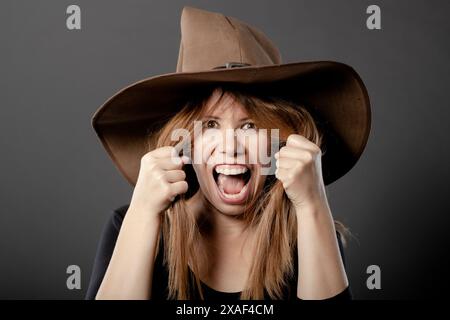 Porträt einer fröhlichen jungen Frau mit einem großen braunen Hut, die Aufregung und Glück mit einem breiten, offenen Jubel zum Ausdruck bringt. Stockfoto