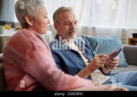 Seitenansicht des Porträts eines modernen Seniorenpaares, das ein Telefon mit mobiler App zusammen nutzt, während es zu Hause auf der Couch sitzt Stockfoto