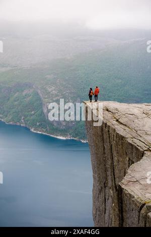 Zwei Individuen stehen am Rande von Preikestolen, einer dramatischen Klippe in Norwegen, mit Blick auf die weite Weite des Fjords darunter. coupe von Männern und Frauen in Preikestolen, Norwegen Stockfoto