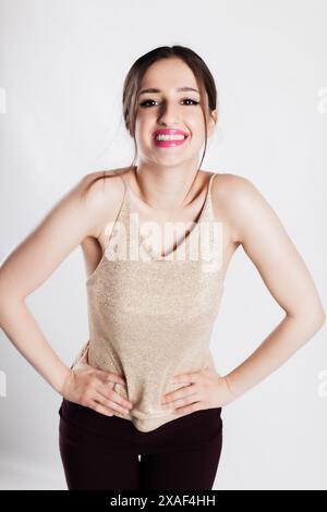 Eine junge Frau steht selbstbewusst in einem glitzernden goldenen Tanktop und lächelt hell vor einem weißen Hintergrund. Stockfoto