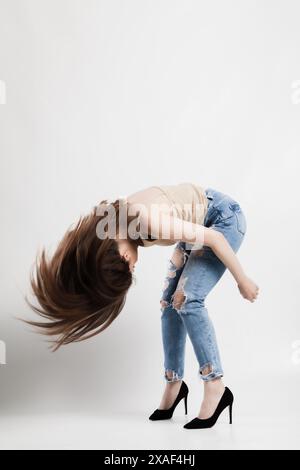 Eine junge Frau fängt mitten in der Bewegung ein, ihre langen Haare fließend, während sie energetisch in stylischen zerrissenen Jeans und schwarzen High Heels auf einem tanzt Stockfoto