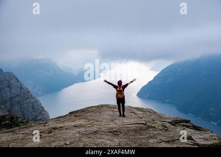 Ein einsamer Wanderer steht mit ausgebreiteten Armen am Rande der berühmten Preikestolen Klippe in Norwegen, mit Blick auf einen atemberaubenden Fjord und die umliegenden Berge. Preikestolen, Norwegen Stockfoto