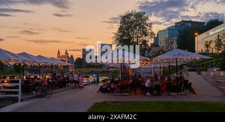 Nachtleben in Vilnius. Die Leute entspannen sich in einem abendlichen Café am Ufer des Neris-Flusses mit zentralem Geschäftsviertel alias Vilnius CBD im Hintergrund Stockfoto