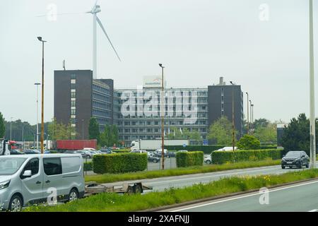 Gent, Belgien - 22. Mai 2023: Ein großes Bürogebäude mit Windkraftanlage im Hintergrund. Autos stehen auf dem Parkplatz vor dem Gebäude. Stockfoto