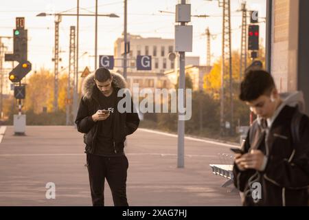 Bild von zwei jungen Männern, die auf einen Zug warten und ihr Smartphone auf dem Bahnsteig des Essener Hbf-Bahnhofs auf der Fahrt im Herbst überprüfen. Stockfoto