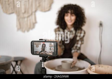 Junge Töpferin, die eine Vase auf einem Töpferrad modelliert, während sie in einem gemütlichen Studio einen Video-Blog mit einem Smartphone aufnimmt. Stockfoto