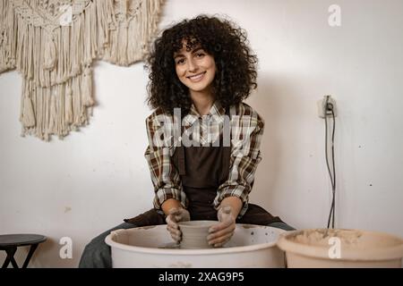 Junge Töpferin, die an Keramikrad arbeitet und Keramikstücke herstellt. Sie lächelt und trägt eine Schürze in einem rustikalen Studio. Stockfoto