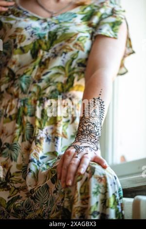 Ein Nahaufnahme-Foto einer Frau, die ein Blumenkleid trägt. Ihr Arm ist mit einem komplizierten Henna-Tattoo verziert, das detaillierte Motive an Hand und Vorderteil zeigt Stockfoto