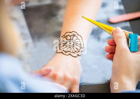 Ein Henna-Künstler wendet ein detailliertes Blumendesign sorgfältig an einem Frauengelenk an, indem er einen mit Henna-Paste gefüllten Kegel verwendet. Die Künstlerhand hält die Stockfoto