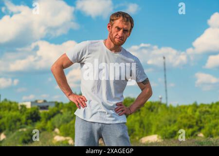 Ein Mann mit weißem T-Shirt steht mit den Händen auf den Hüften, während er sich während eines Workouts im Freien in einem Park an einem sonnigen Tag erwärmt. Stockfoto