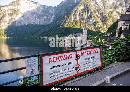 18. Mai 2024, Österreich, Hallstatt: Die katholische Pfarrkirche Hallstatt und die evangelische Pfarrkirche Hallstatt sind am 18.05.2024 in Hallstatt in Oberösterreich vor dem Alpenpanorama über den Hallstätter See zu sehen. Im Vordergrund steht ein Banner auf einem Zaun in zwei Sprachen: „Achtung - Wir leben hier! Bitte genießen Sie die schöne Aussicht in Ruhe, ohne lautes Schreien oder Musik.“ Daneben weist ein kleines Schild auf die „No Drone Zone“ hin. Der Aussichtspunkt am Hallstätter See ist zu einem echten „Insta-Hotspot“ geworden, der jeden Tag unzählige Touristen anzieht. Foto: Matthias Balk/dpa Stockfoto