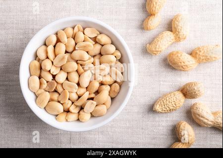 Geröstete und gesalzene Erdnüsse in einer weißen Schüssel auf Leinenstoff. Verzehrfertige Snacks, hergestellt aus Früchten der Arachis hypogaea, auch bekannt als Erdnuss. Stockfoto