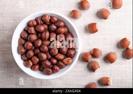 Haselnüsse in einer weißen Schüssel auf Leinenstoff. Ganze, getrocknete und geschälte Nüsse von Corylus avellana, rechts in der Schale. Bereit zum Essen als Snack. Stockfoto