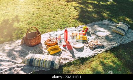 Picknickdecke mit gekochtem Mais, Früchten, Kuchen, einer Flasche Saft, Korb, Stillleben im Sommerpark der Stadt. Keine Menschen. Stockfoto