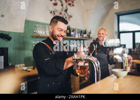 Stilvoller bärtiger Barkeeper in schwarzer Uniform, lächelnd in die Kamera, Bierzapfen an der Bar und Kellner mit Tablett. Erfolgreiche Teamarbeit, Freundlichkeit Stockfoto