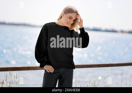 Mockup eines schwarzen Sweatshirts auf einem Mädchen vor dem Hintergrund eines Flusses, Pier, Vorderansicht der Kleidung. Modische, langärmelige Damenbekleidung Stockfoto