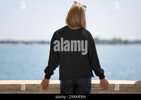 Vorlage eines schwarzen Sweatshirts auf einem blonden Mädchen bei sonnigem Wetter auf dem Pier, auf dem Hintergrund eines Flusses, Rückansicht, für Werbung. Modell von Stockfoto