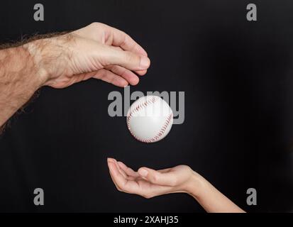 Mann Hand wirft einen Baseball in eine Frauenhand auf schwarzem Hintergrund Stockfoto