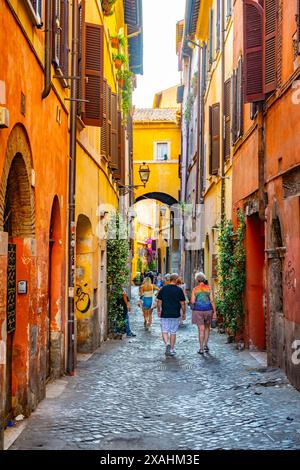 Touristen spazieren durch eine enge, kopfsteingepflasterte Gasse in Rom, Italien. Die Gebäude sind in hellen Farben gehalten und mit Pflanzen und Blumen geschmückt. Die Gasse führt zu einem Bogengang, der einen Blick auf die Stadt dahinter bietet. Stockfoto