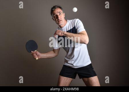 Porträt des konzentrierten Tischtennisspielers, der einen orangen Ball auf grauem, isoliertem Hintergrund trifft. Stockfoto