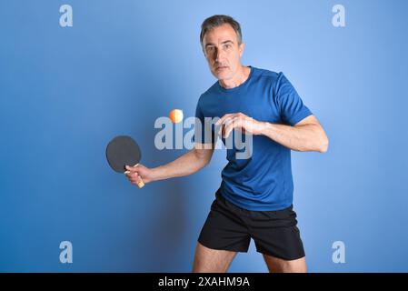 Porträt des konzentrierten Tischtennisspielers, der einen orangen Ball auf blauem isoliertem Hintergrund trifft. Stockfoto