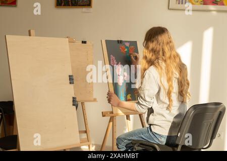 Rückansicht einer kaukasischen, langhaarigen Künstlerin auf Leinwand in einem sonnendurchfluteten Atelier, umgeben von leeren Leinwänden. Stockfoto