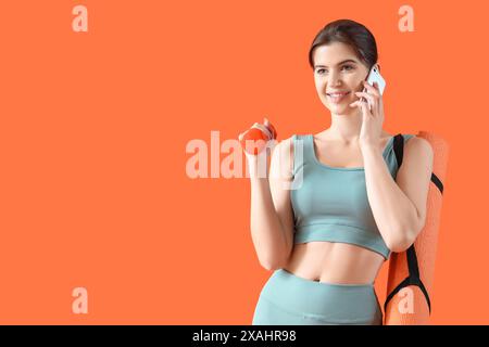 Junge Frau in Sportbekleidung mit Kurzhantel und Fitnessmatte, die mit Handy auf orangem Hintergrund spricht Stockfoto