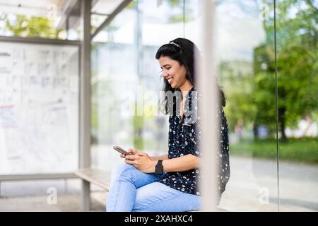 Junge Frau, die an der Bushaltestelle in der Stadt ihr Smartphone benutzt Stockfoto