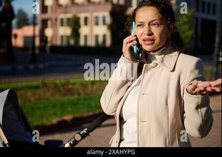 Frustrierte Frau, die mit ihrem Telefon telefoniert, während sie mit einem Kinderwagen in einem sonnigen Stadtgebiet läuft. Sie scheint etwas Wichtiges zu besprechen, sie Stockfoto