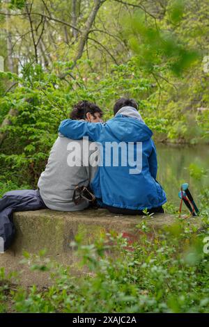 Vertikales Bild der Rückansicht von zwei Personen, die auf einem Felsen in der Nähe eines umarmten Flusses sitzen Stockfoto